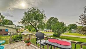 Um deck de madeira com mesa e cadeiras, um vaso de planta e vista para uma área residencial com casas e árvores.