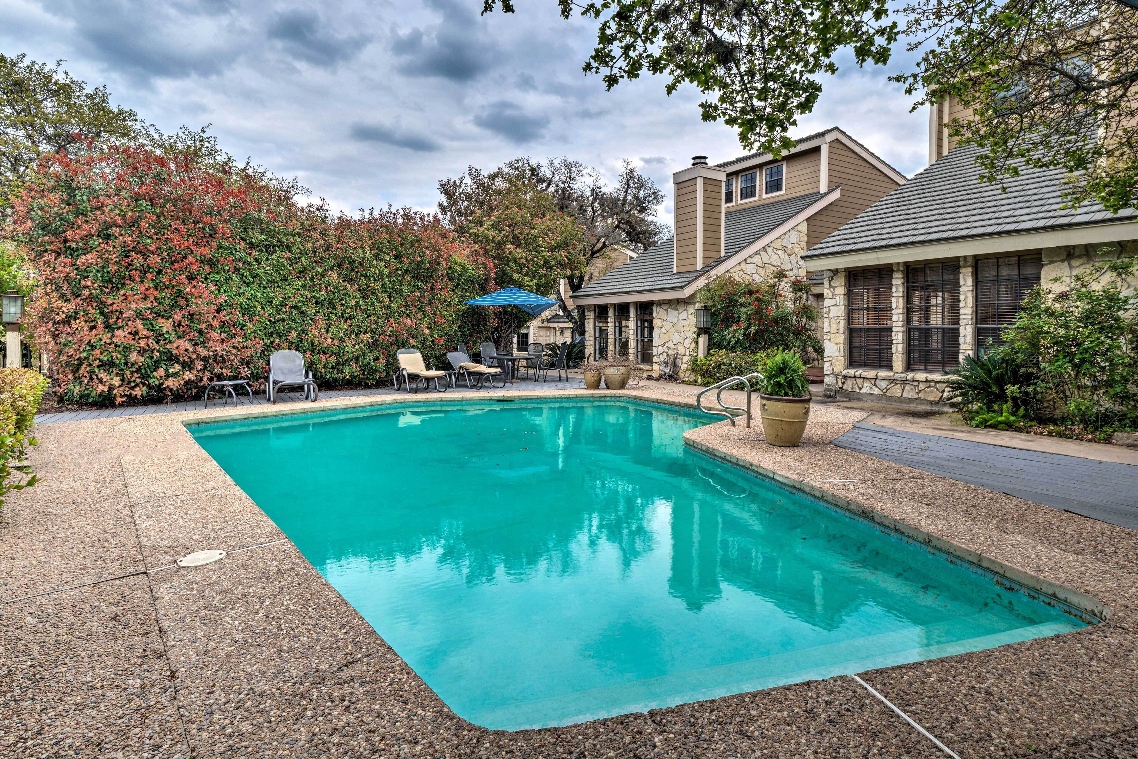 A rectangular swimming pool with a pebble-lined edge, surrounded by a patio area with outdoor furniture and a stone building in the background.