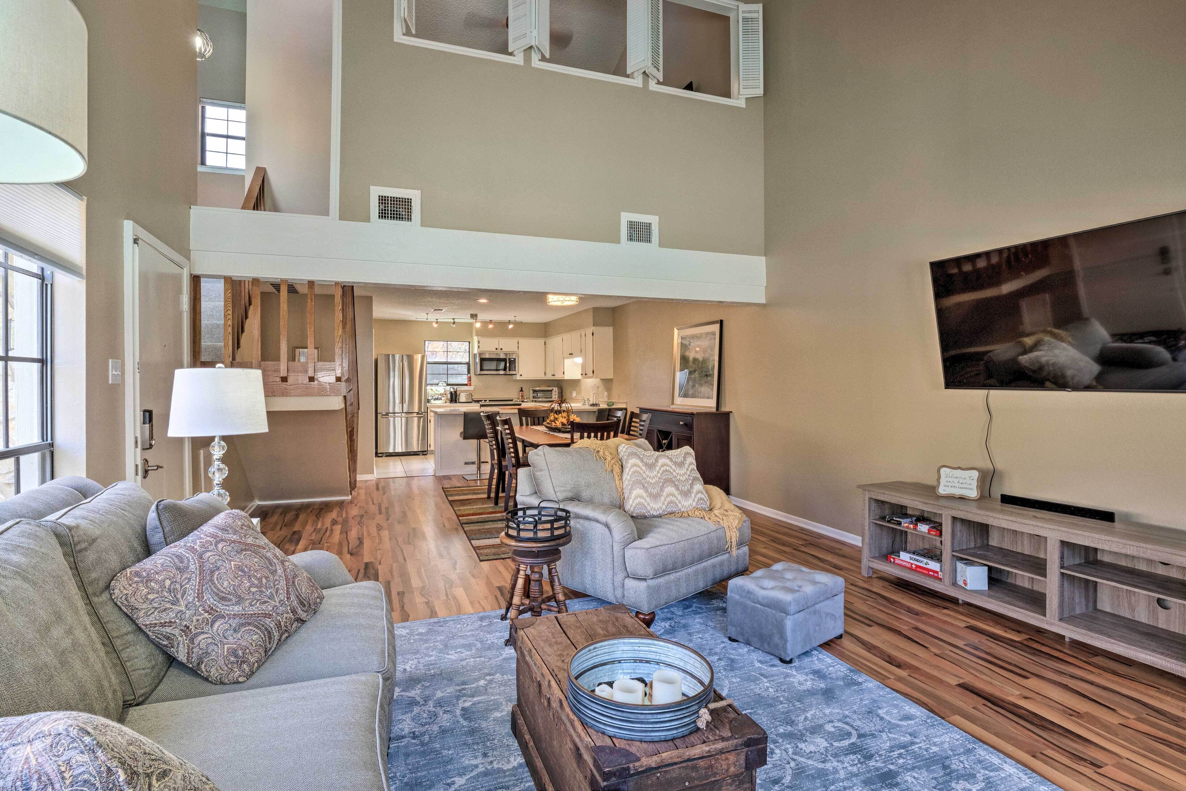 A spacious living area with a high ceiling, a kitchen in the background, and a large TV mounted on the wall.