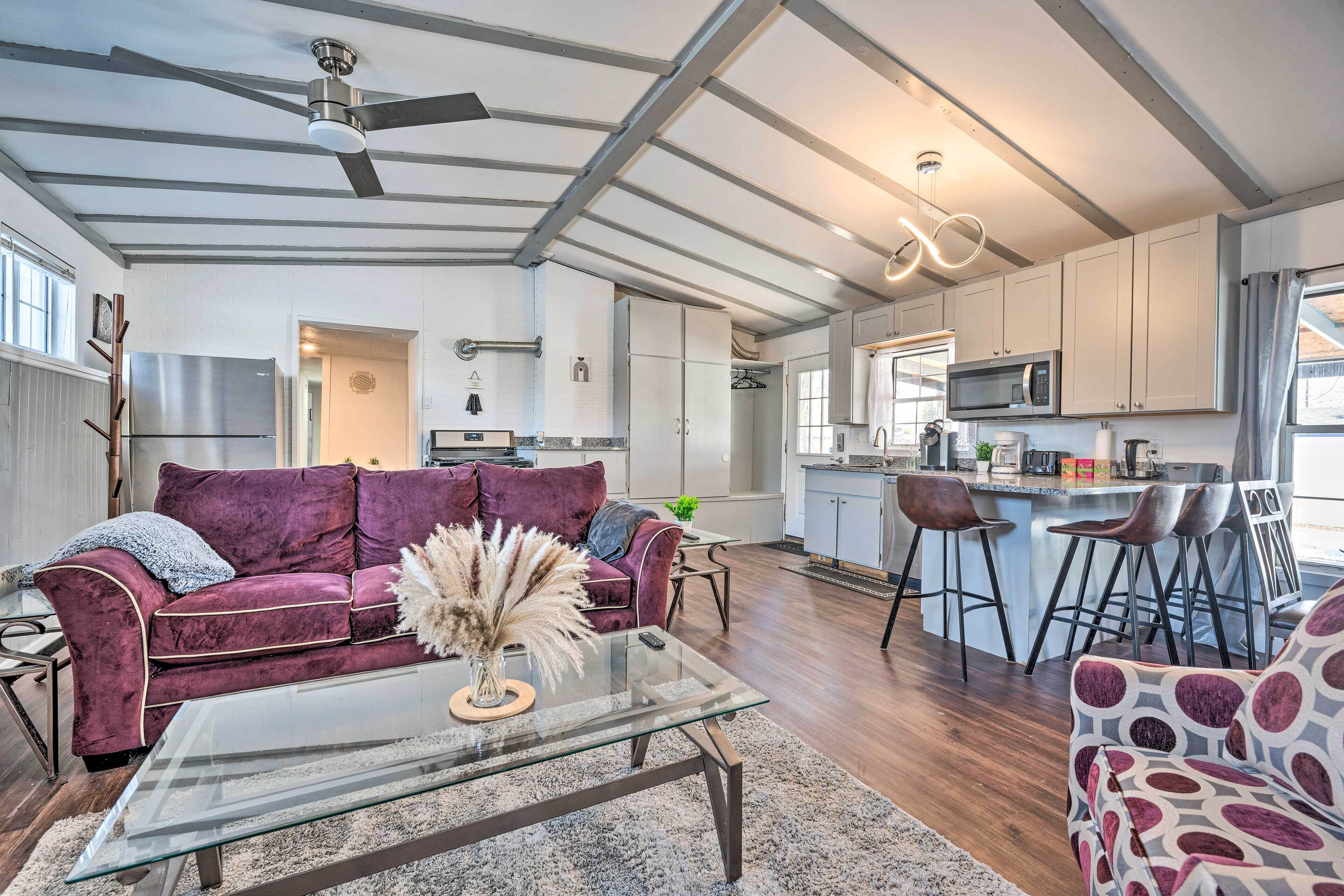A modern living room with a glass coffee table, a purple sofa, and a kitchen area with a bar and seating.