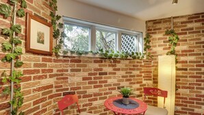 Una habitación con pared de ladrillo, una ventana, un cuadro enmarcado, una mesita con una planta y dos sillas.