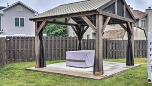 Un quiosco de madera con una mesa cubierta con tela blanca, ubicado en un patio de concreto en un jardín trasero con una cerca de madera y casas al fondo.