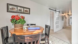 A dining area with a round wooden table, four chairs, a basket of red flowers, a notebook, and a blue book. There are framed pictures on the wall above the table.