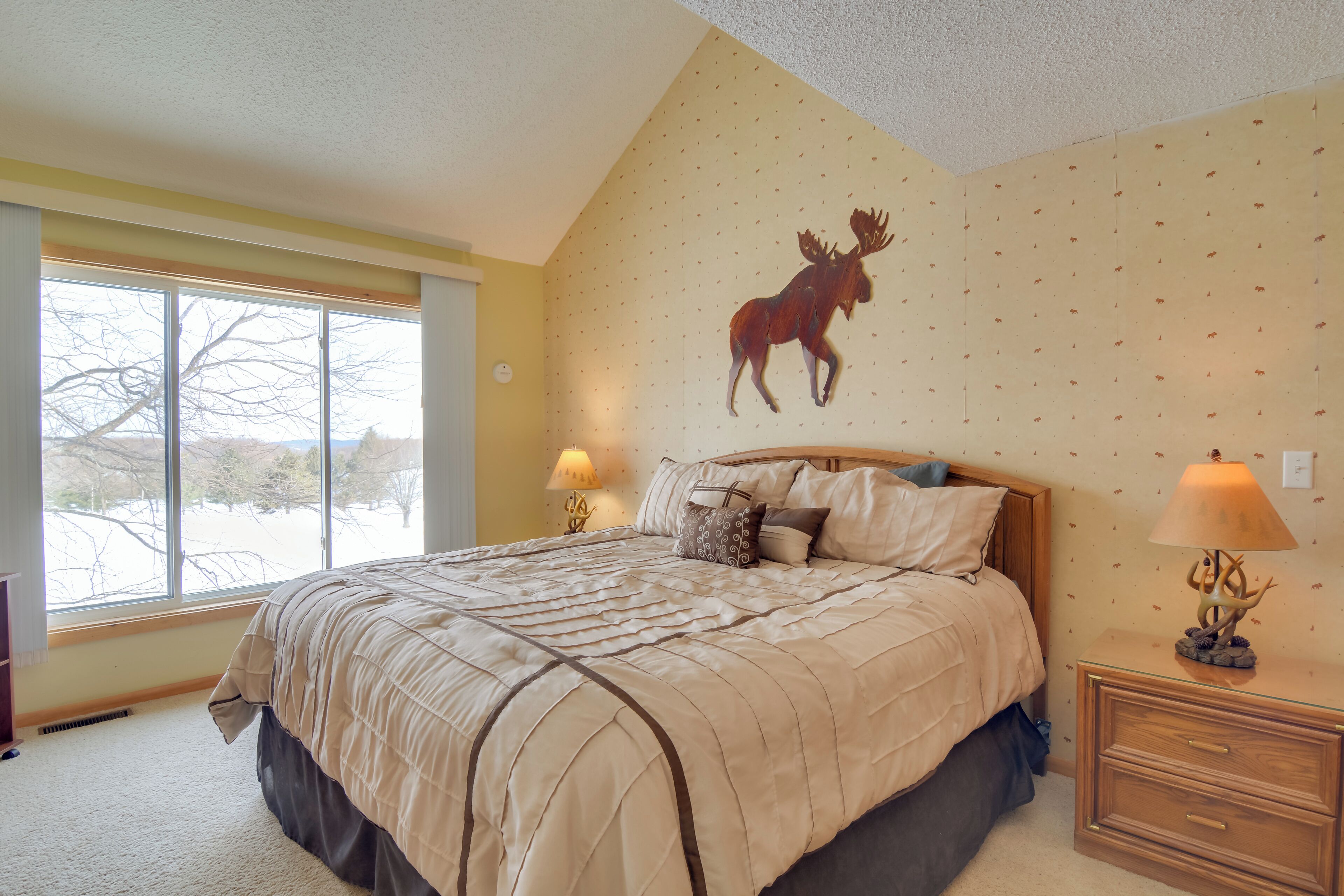 A bedroom with a large bed, a wooden headboard, two bedside tables with lamps, a wall mural of a moose, and a window with a view of trees.