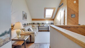 A cozy loft-style room with a wooden ceiling, a skylight, a sofa, a table, and a small kitchen area.