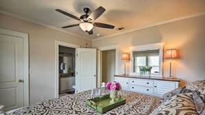 A bedroom with a patterned bedspread, a ceiling fan, a dresser with a mirror, and a nightstand with a lamp. - Inviting Lubbock Home > 1 Mi to Downtown (Lubbock)