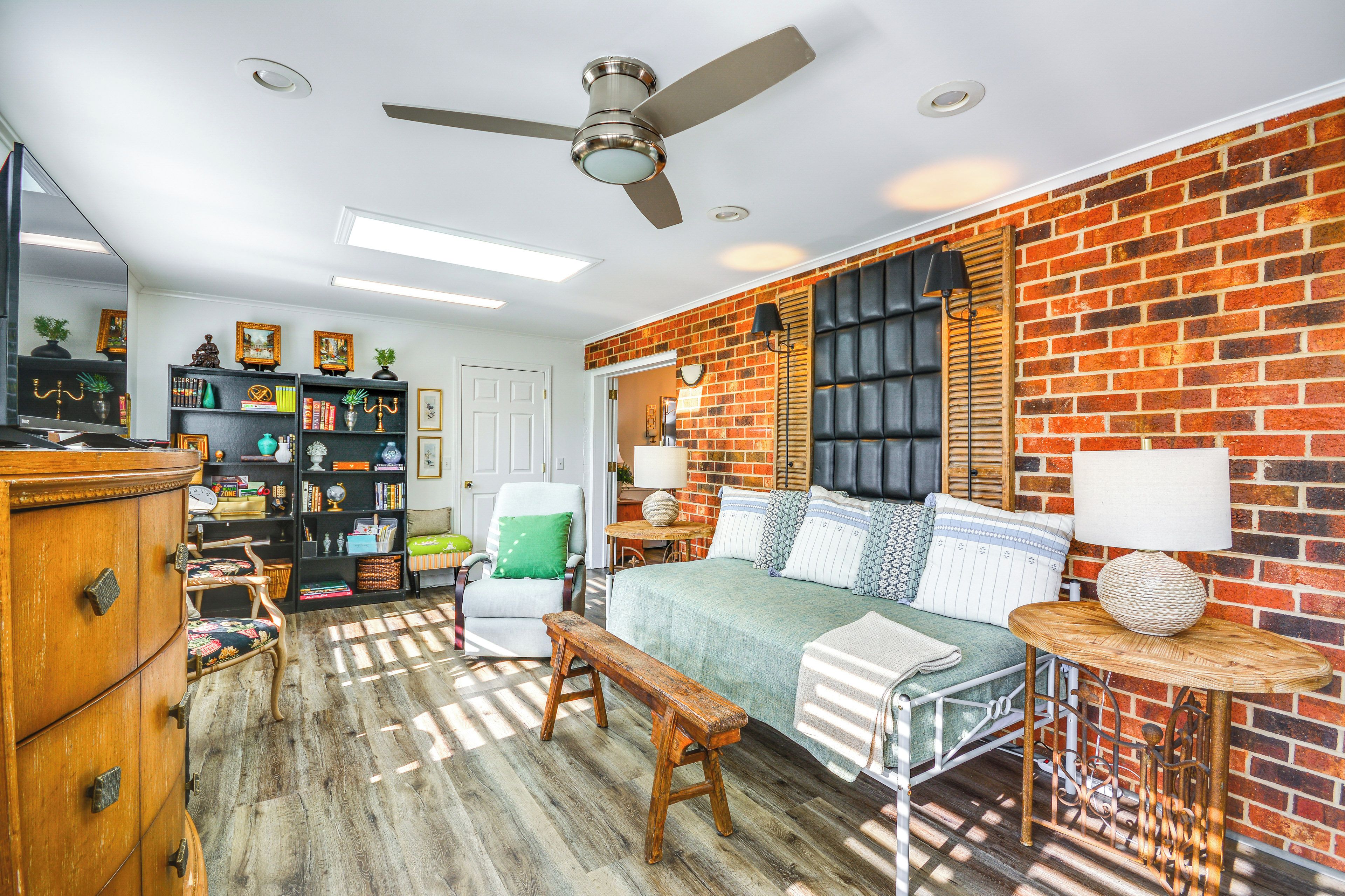 A room with a brick wall, a bed with pillows, a bench, a chair, a bookshelf, and a ceiling fan.
