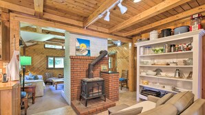 A cozy living room with a fireplace, a sofa, and a bookshelf filled with kitchenware. - Boothbay Harbor Cabin w/ Spacious Deck & Yard! (Boothbay Harbor)