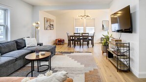 A modern living room with a grey sofa, a coffee table, and a dining area with a chandelier. - Hudson Valley Vacation Rental: Near Hiking (Beacon)