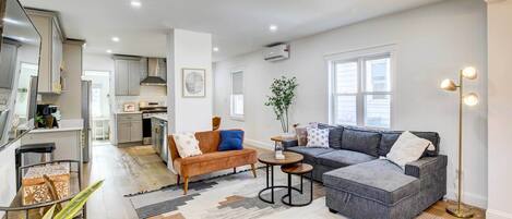 A modern living room with a grey sectional sofa, a brown sofa, and a coffee table. There is a kitchen in the background with white cabinets and a window.