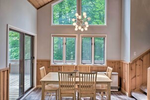 Comedor con mesa y sillas de madera, ventanas amplias con vistas a árboles y una lámpara de araña.