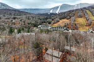 Un edificio grande con múltiples ventanas y una entrada destacada, rodeado de árboles y un paisaje montañoso.