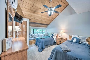 A bedroom with a sloped ceiling, a ceiling fan, a bed with a blue comforter, a bedside table with a lamp, and a window with a view of greenery. - Village of Loon Condo in Lincoln w/ Pool Access (Lincoln)