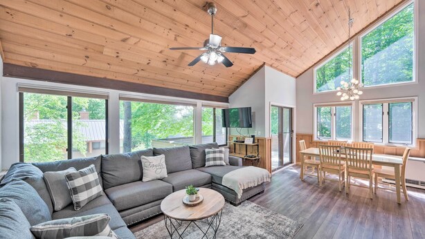 A spacious living room with a large sectional sofa, a dining table with chairs, a ceiling fan, and large windows offering a view of the outdoors. - Village of Loon Condo in Lincoln w/ Pool Access (Lincoln)