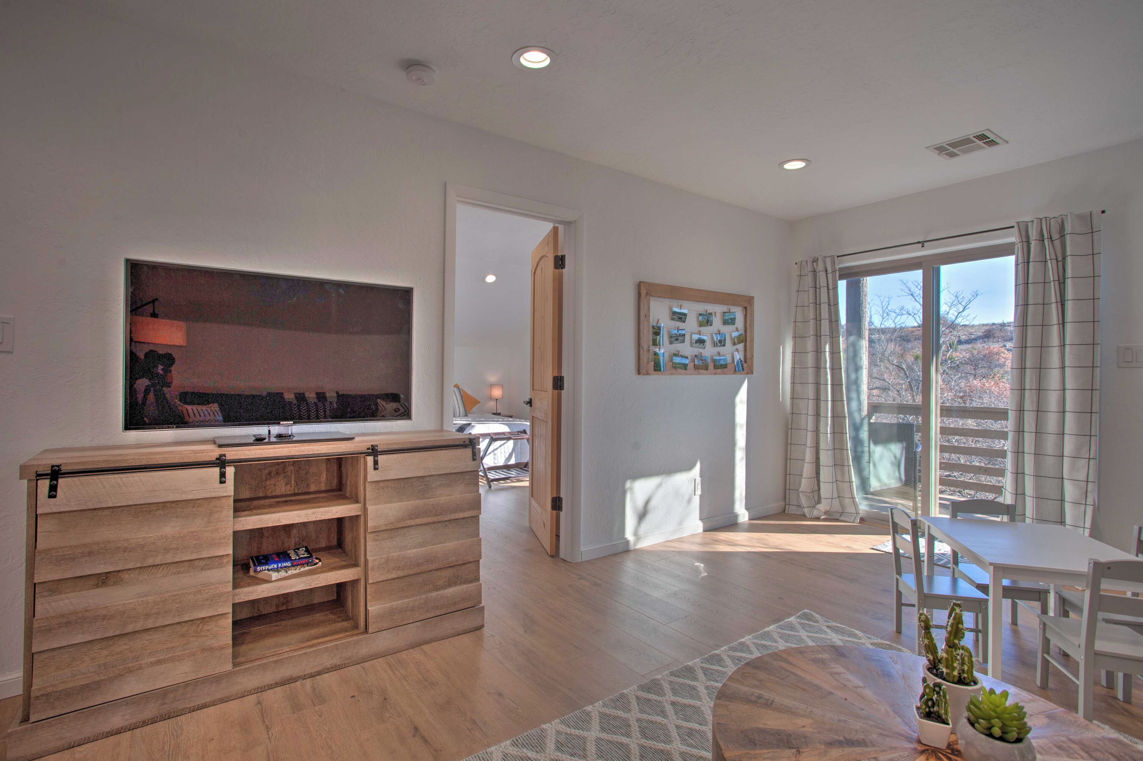 A modern living room with a wooden entertainment unit, a dining area with white furniture, and a large window with curtains.