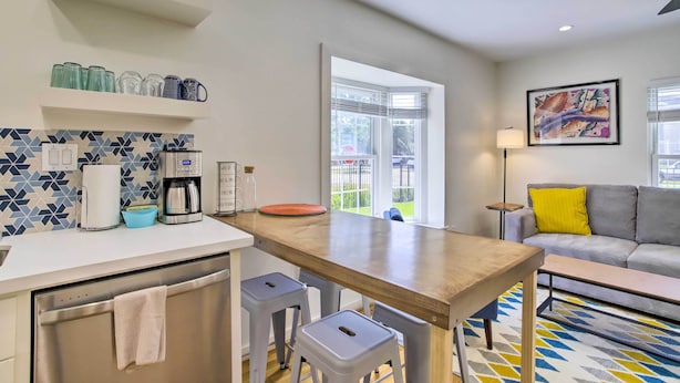 Cozinha moderna com mesa de madeira, banquetas e máquina de café. Há um sofá e um quadro na sala de estar.