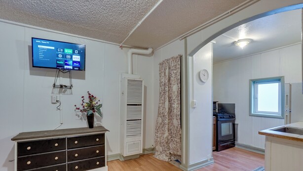 A living room with a flat-screen TV, a wooden dresser, a patterned rug, and a kitchen area with a stove and refrigerator.