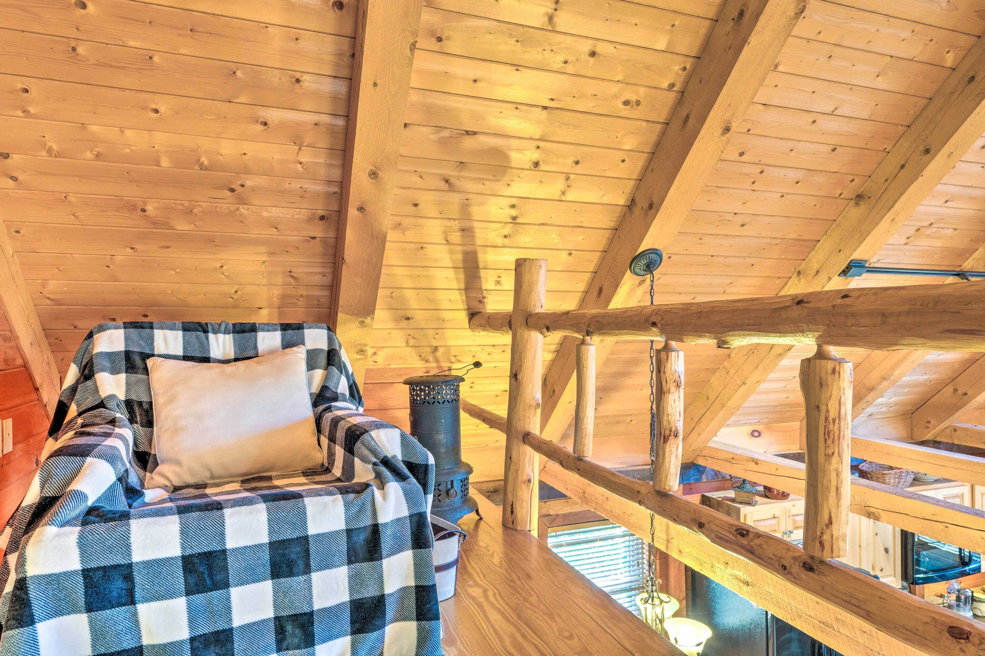 A cozy wooden cabin interior with a checkered blanket on a bed, a rustic wooden ceiling, and a wooden railing overlooking the cabin.