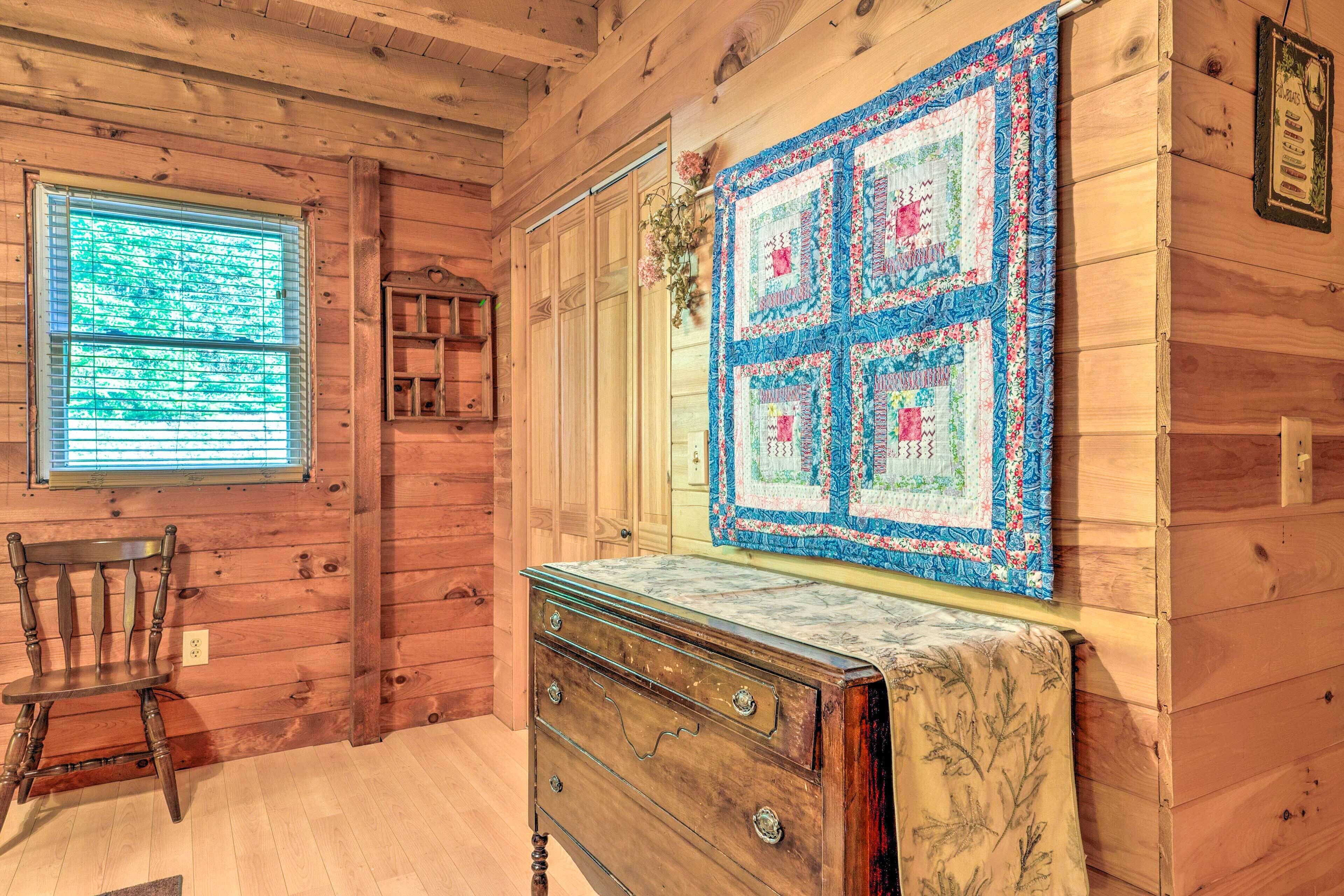 Interior de una cabaña de madera con un aparador de madera, una silla, un colcha y una ventana con persianas.