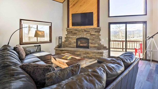 A modern living room with a stone fireplace, leather sofa, and large windows offering views of the outdoors. - Spacious Fairplay Cabin w/ Views ~ 21 Mi to Breck! (Alma)