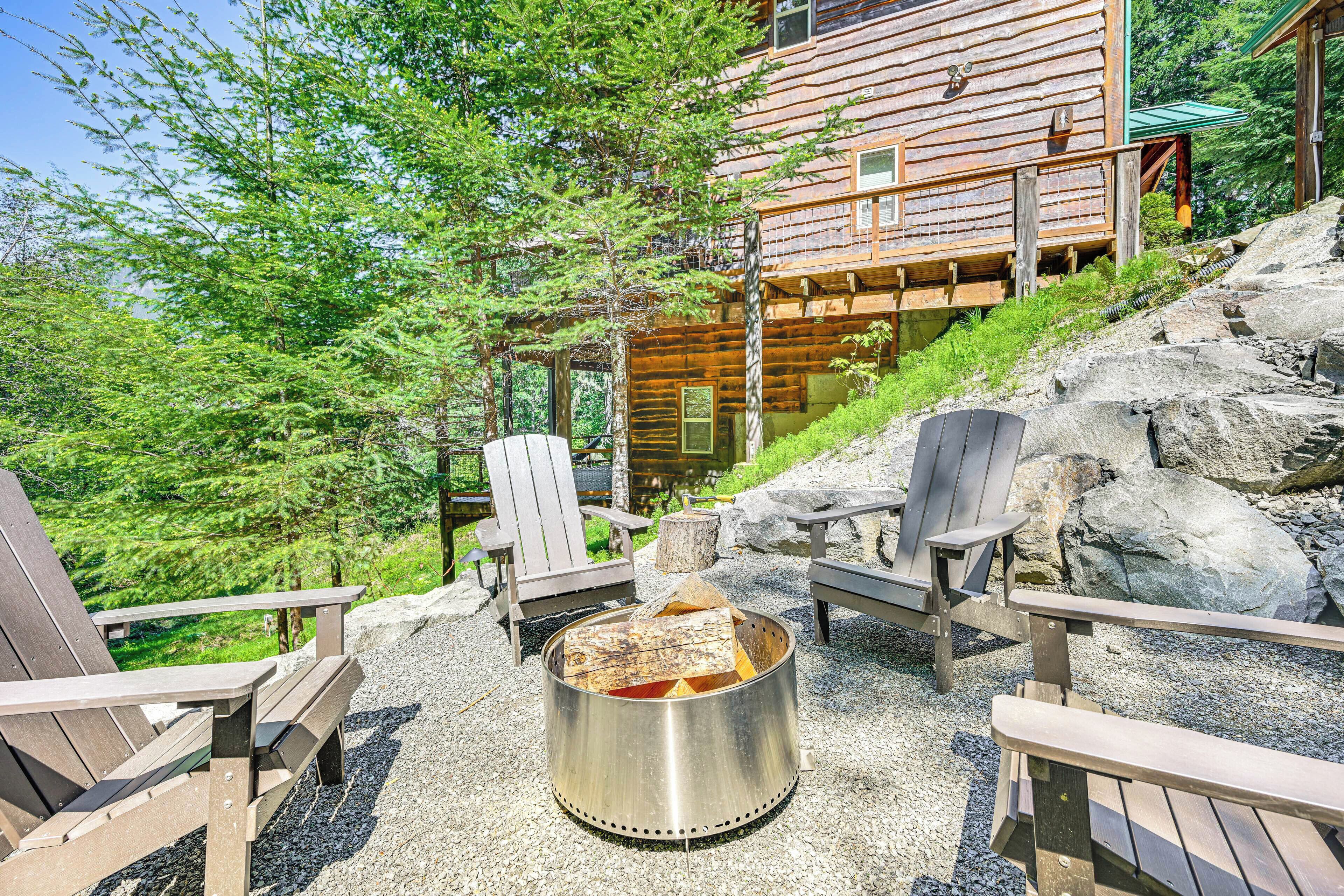 Uma cabana de toras com uma lareira cercada por cadeiras Adirondack, com árvores e um morro rochoso ao fundo.