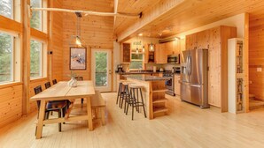 Una cabaña de madera espaciosa con una gran mesa de comedor, taburetes de bar, una zona de cocina con electrodomésticos de acero inoxidable y ventanas amplias que ofrecen vistas al exterior.