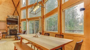 Un interior de madera espacioso con una gran mesa de comedor, chimenea de piedra y amplias ventanas con vistas al bosque.
