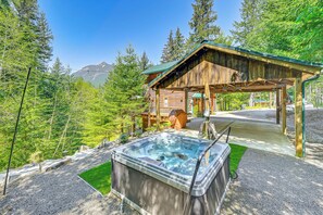 Une cabane en bois dotée d’un bain à remous et d’une terrasse couverte, entourée d’arbres et avec une montagne en toile de fond.