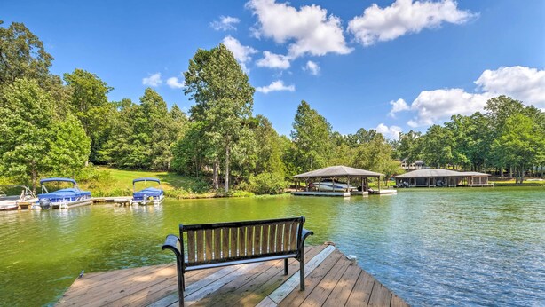 A wooden dock with a bench, a calm lake, boats, and a pavilion in a wooded area.