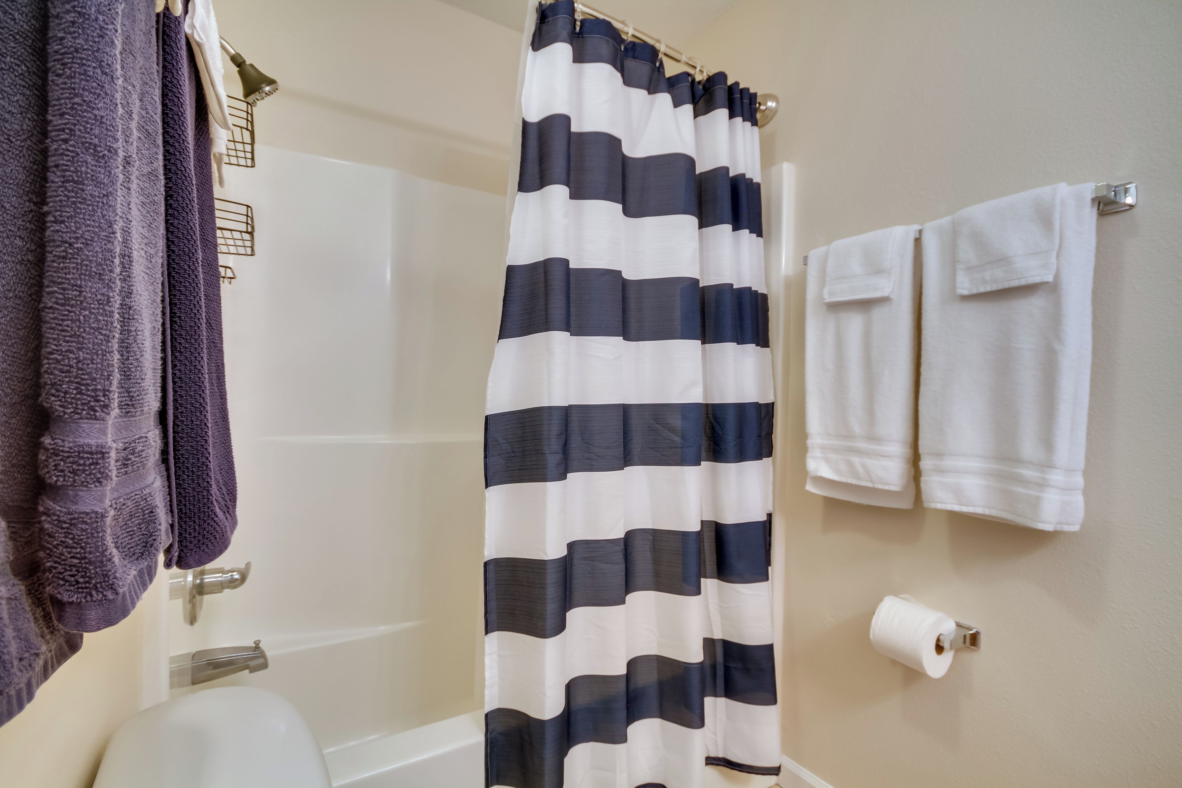 Ein Badezimmer mit gestreiftem Duschvorhang, einer weißen Badewanne, zwei weißen Handtüchern auf einem Haken und einem Toilettenpapierhalter.