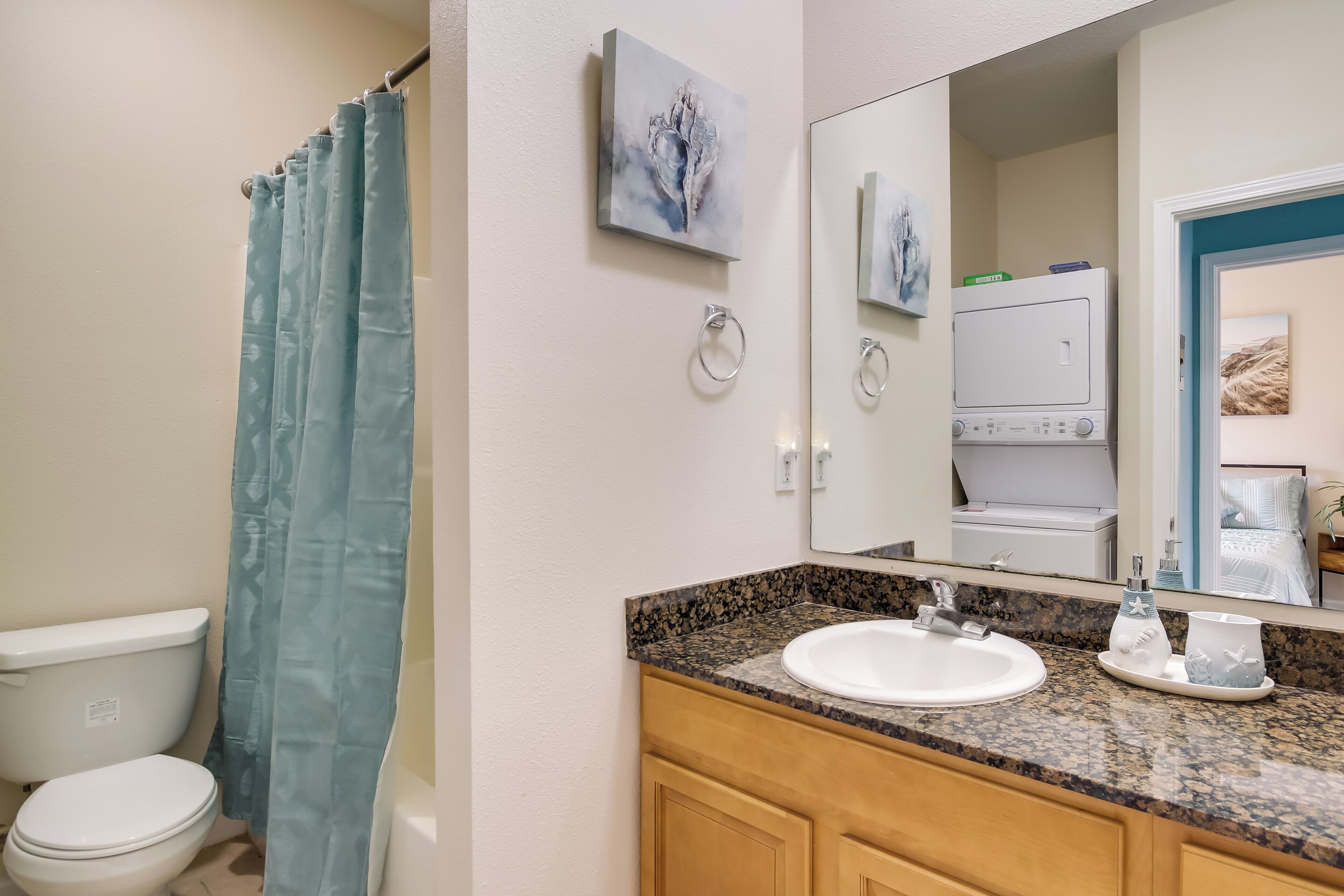 A bathroom with a white toilet, a blue shower curtain, a granite countertop with a sink, and a mirror reflecting a shower area.