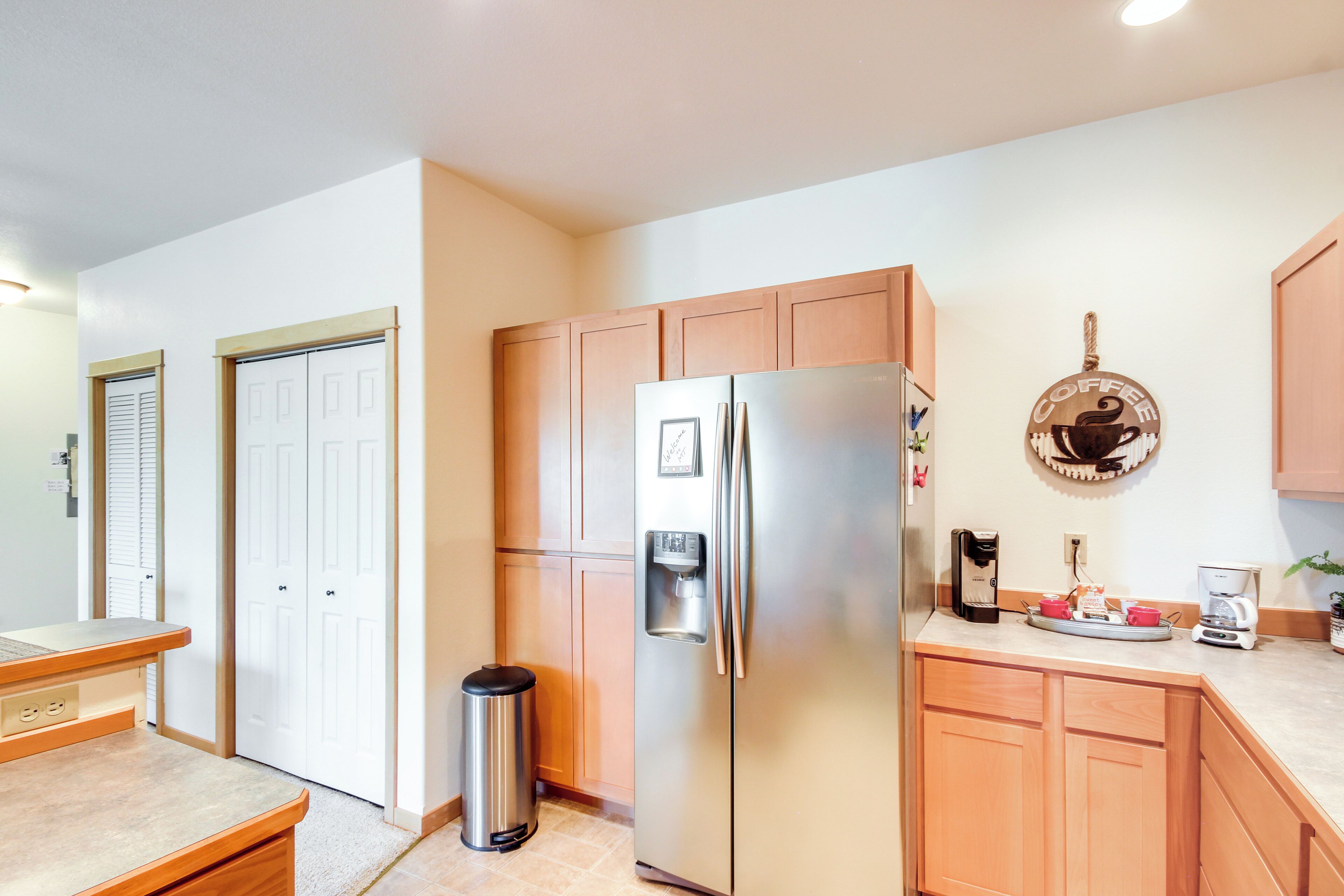 A modern kitchen with a stainless steel refrigerator, wooden cabinets, a coffee maker, and a trash can.