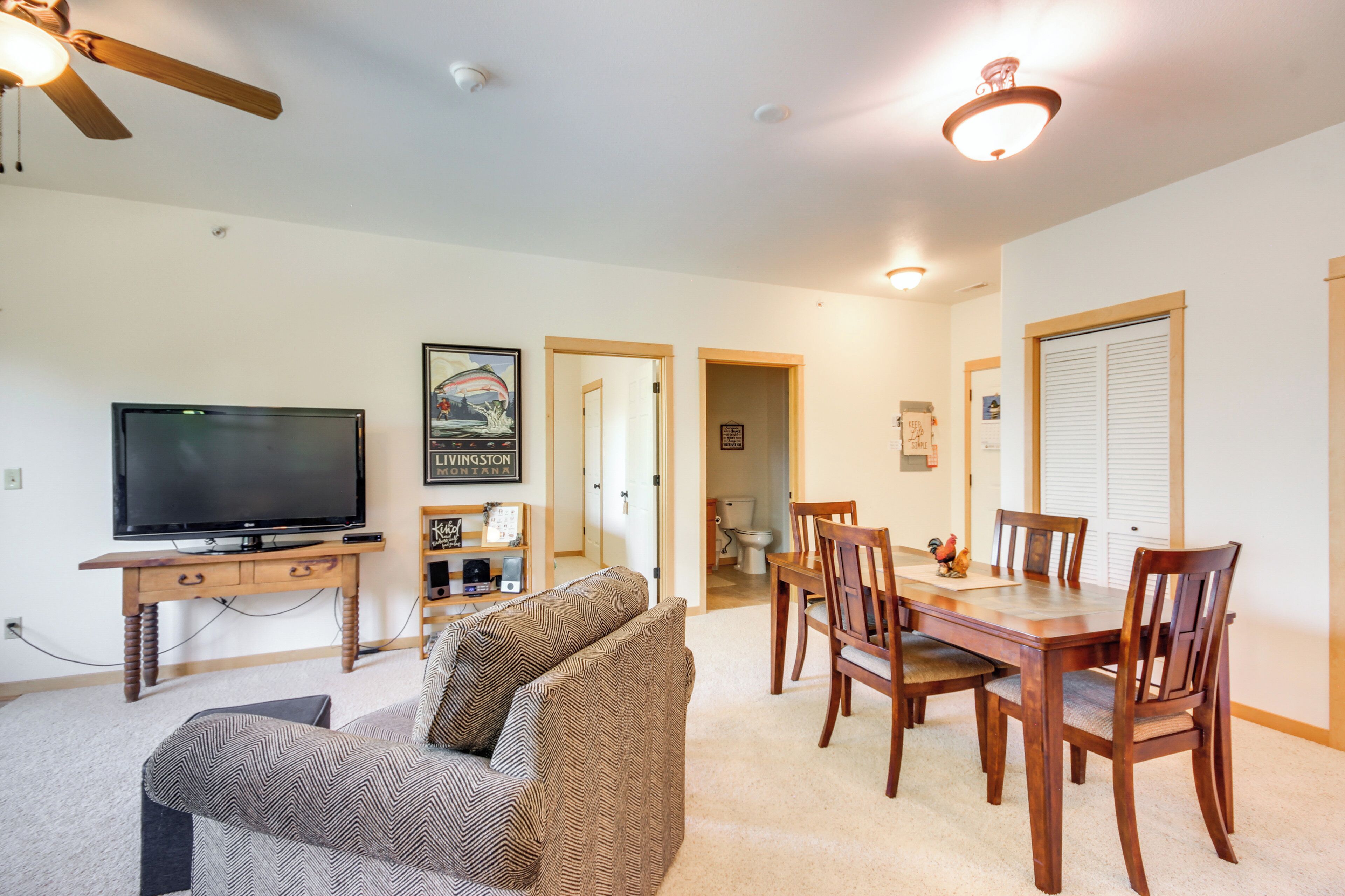 A living room with a television, sofa, wooden dining table, and chairs. There is a ceiling fan and a light fixture.