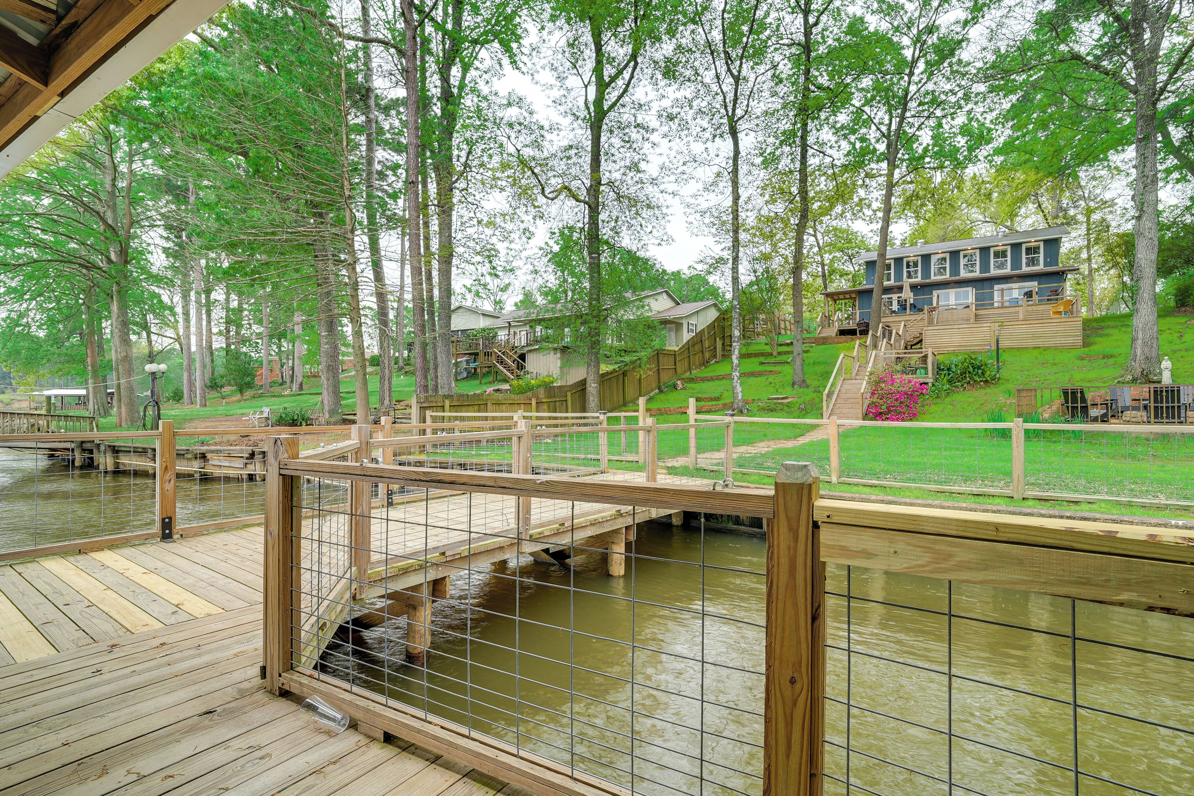 A wooden deck with a bridge over a water body, surrounded by trees and a house in the background.