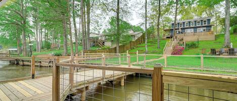 A wooden deck with a bridge over a water body, surrounded by trees and a house in the background.