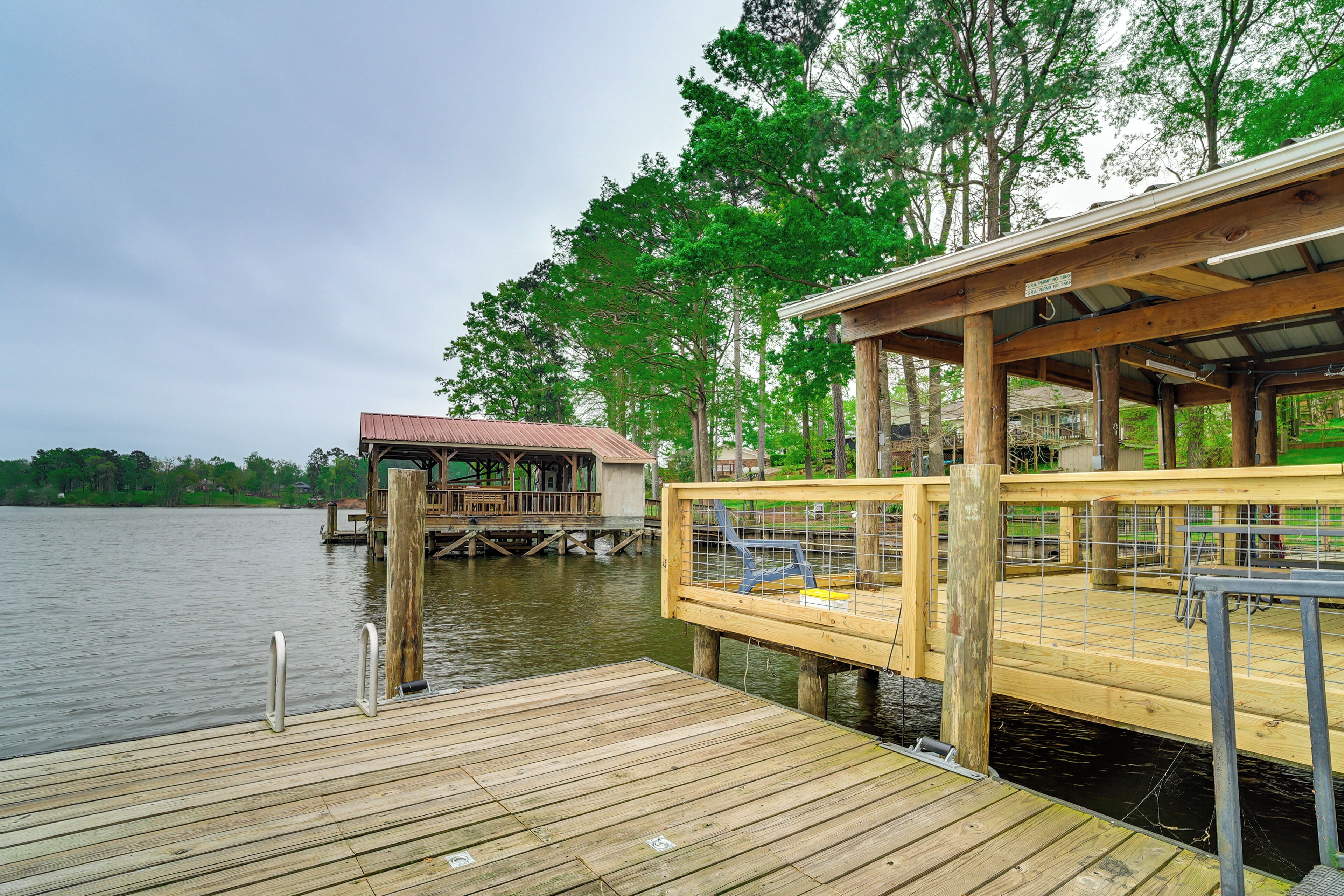A wooden dock extending into a calm lake, with a covered seating area and a small building in the background.