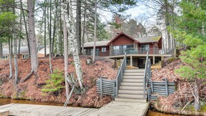 A cabin with a wooden deck leading to a dock over a river, surrounded by trees.