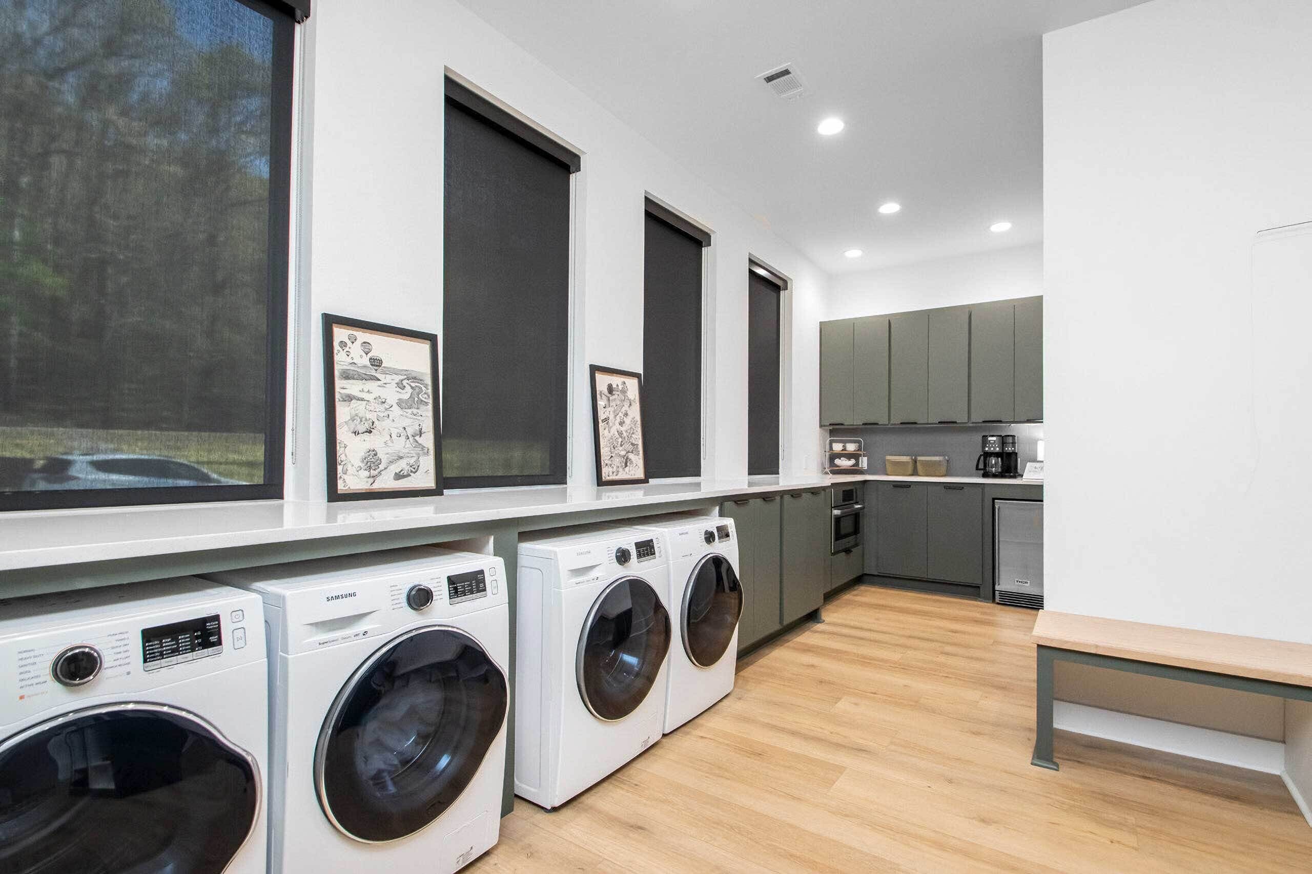 A modern laundry room with four Samsung washing machines, a bench, and framed artwork on the wall.