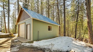 Una cabaña pequeña de un solo piso con techo metálico y fachada verde, rodeada por un área boscosa con nieve en el suelo.