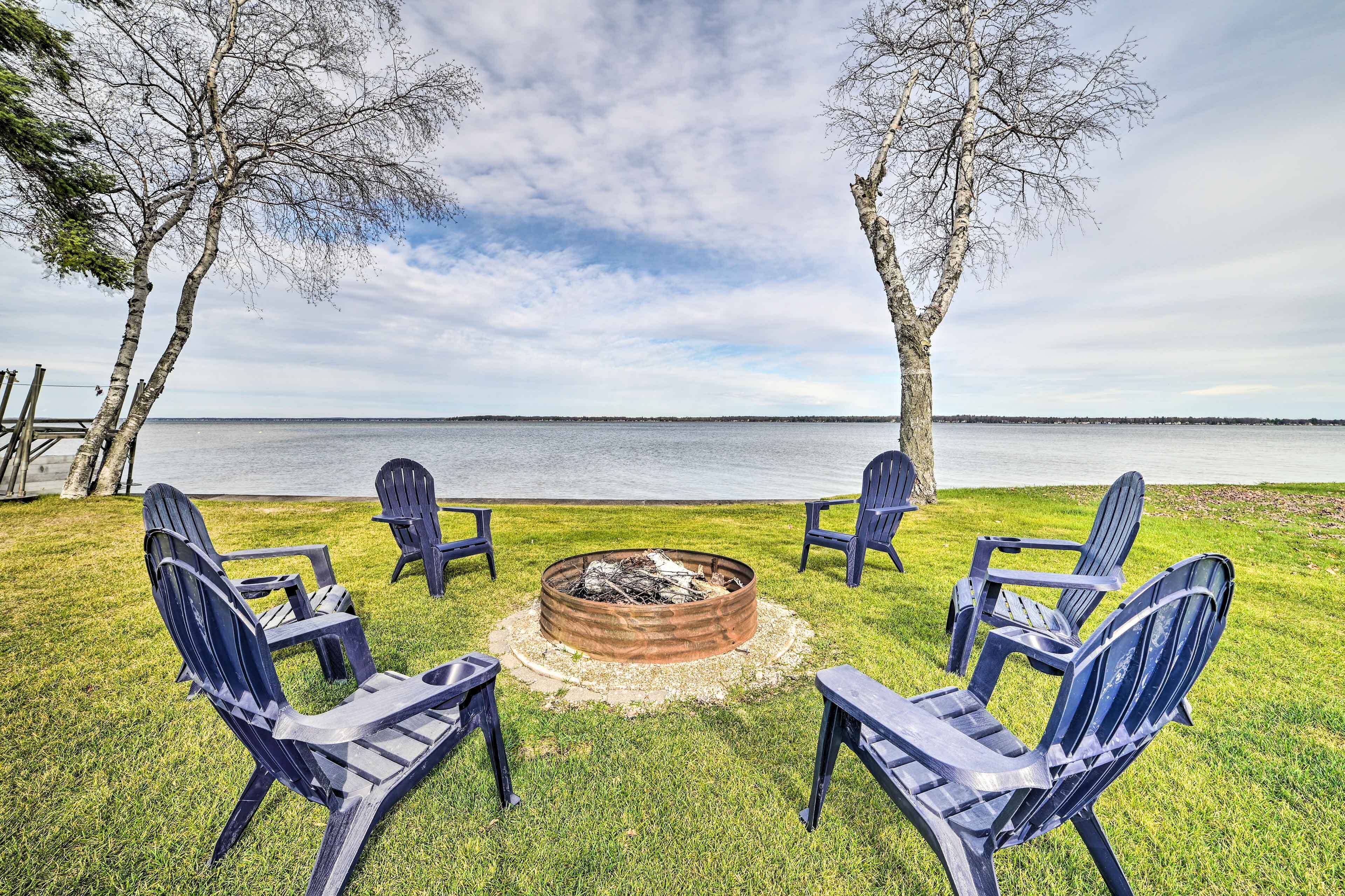 A fire pit surrounded by Adirondack chairs on a grassy area with a lake view.