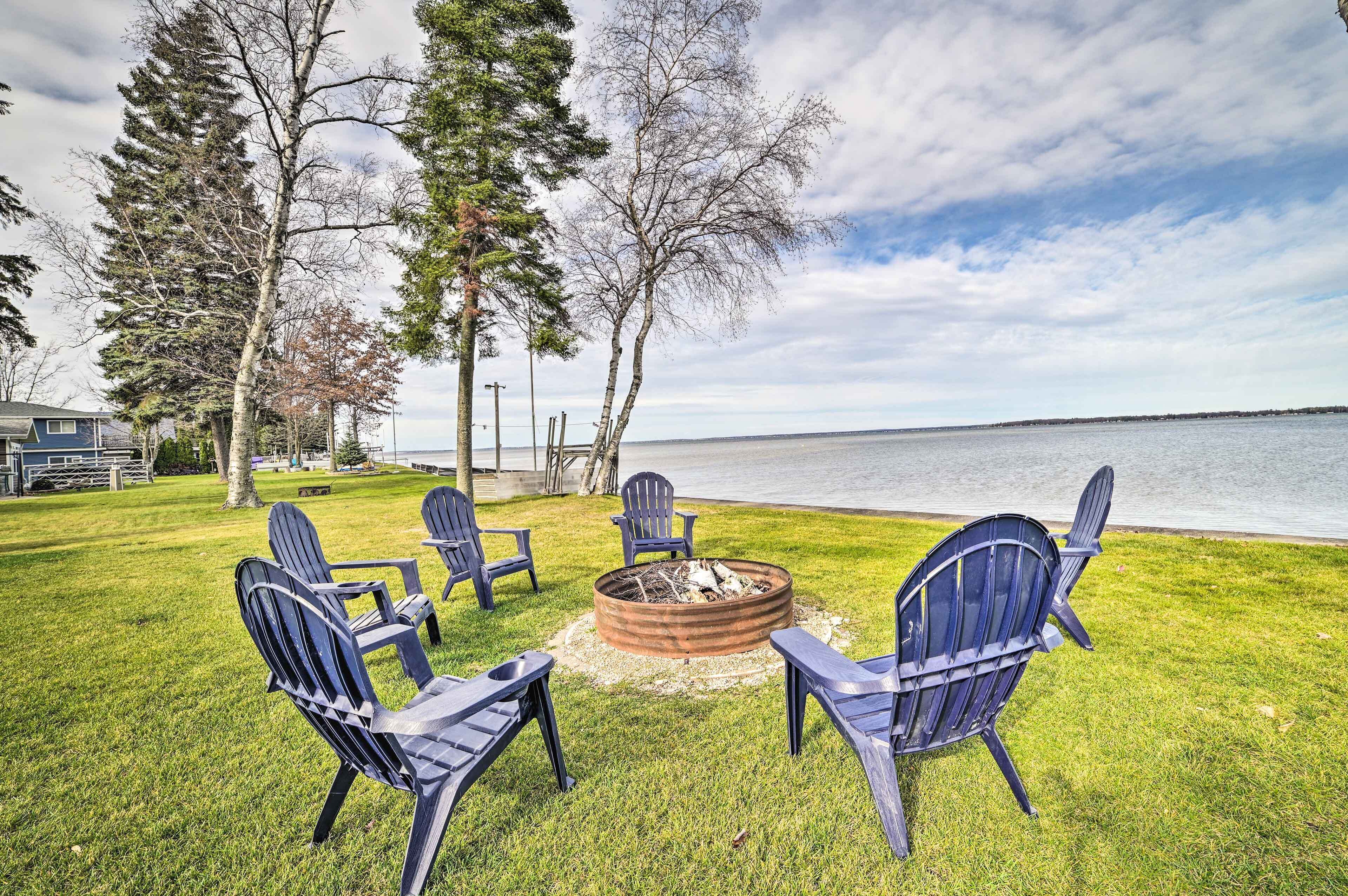 A grassy outdoor area with a fire pit and Adirondack chairs facing a body of water.