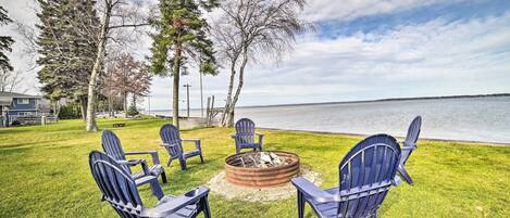 A grassy outdoor area with a fire pit and Adirondack chairs facing a body of water.