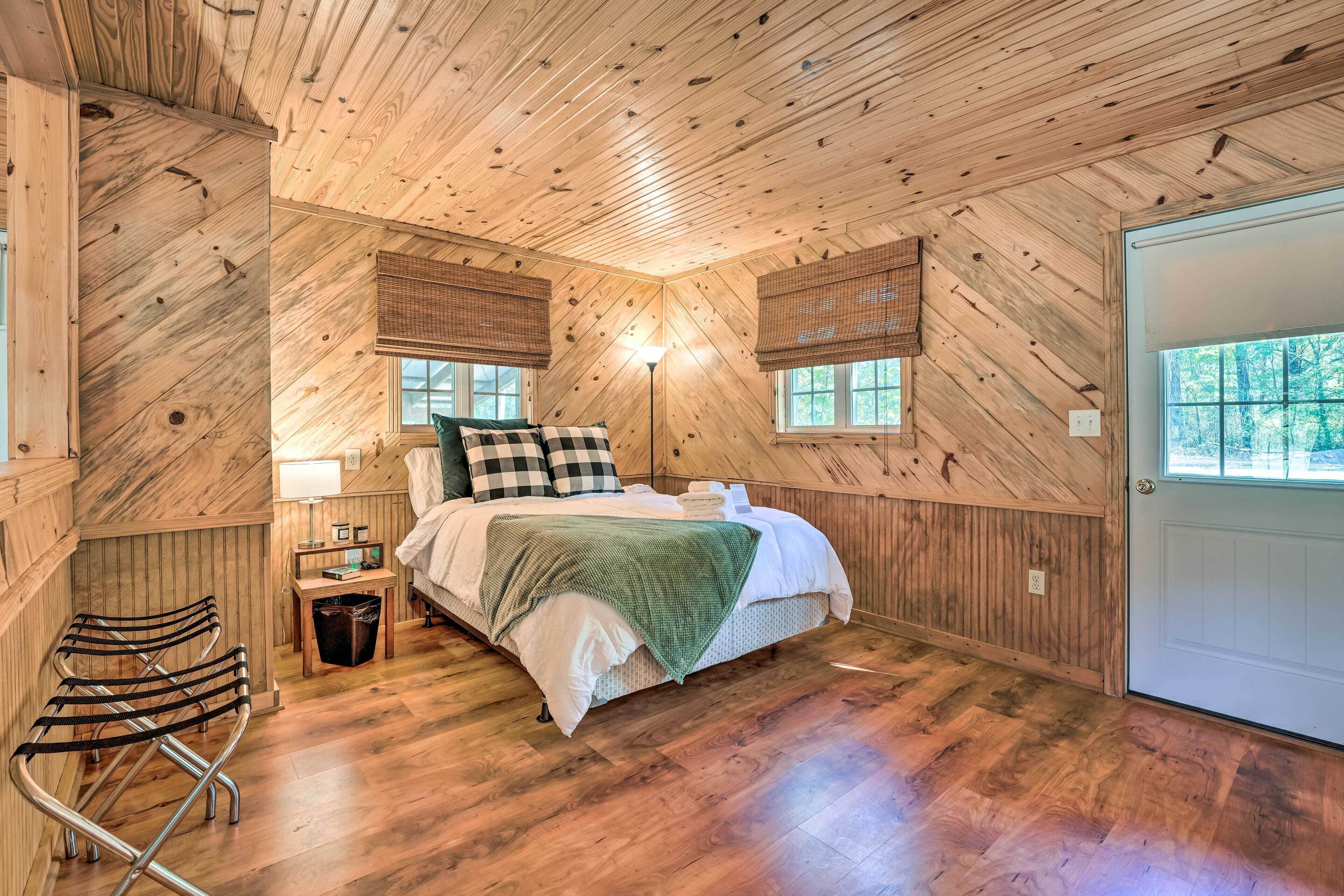 Ein gemütliches Schlafzimmer mit Holzwänden und -decke, einem Bett mit grüner Decke, einem Nachttisch mit Lampe und einem Fenster mit Blick auf Bäume.
