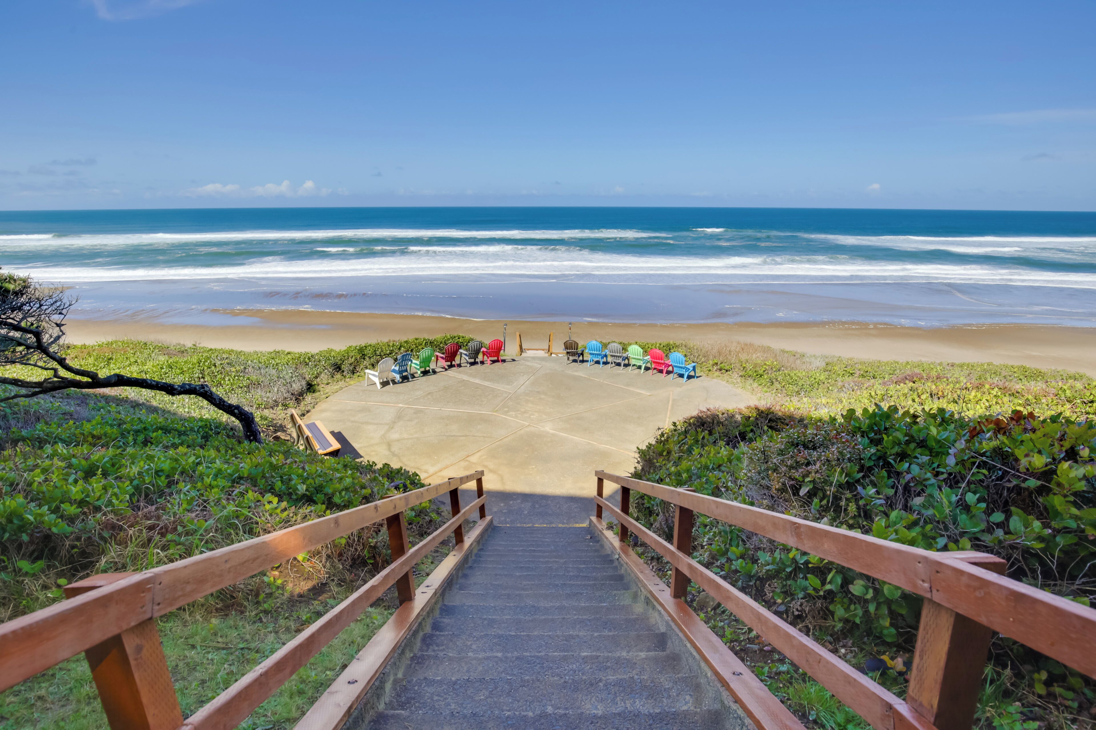Een houten trap die naar een strand leidt, met kleurrijke stoelen in een halve cirkel gerangschikt.