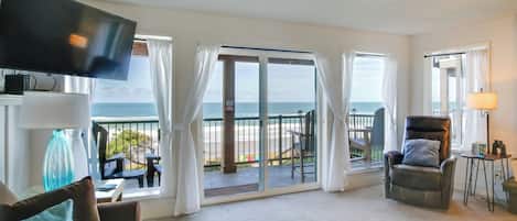 A spacious living room with a large TV, a sofa, a coffee table, and a view of the beach through a sliding glass door.