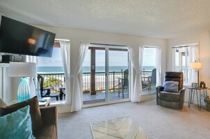 A spacious living room with a large TV, a sofa, a coffee table, and a view of the beach through a sliding glass door.