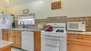Une cuisine avec des meubles de rangement en bois, un lave-vaisselle, une cuisinière, un four à micro-ondes et une fenêtre agrémentée d’un rideau à motifs.