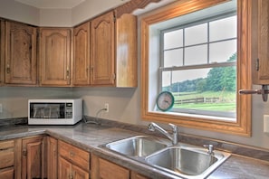 A kitchen with wooden cabinets, a stainless steel sink, a microwave, and a window with a view of greenery.