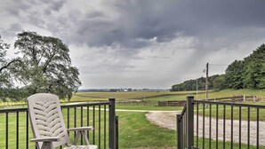 Eine Holzterrasse mit einem weißen Stuhl, einem Metallgeländer und Blick auf ein grasbewachsenes Feld und Bäume unter einem bewölkten Himmel.