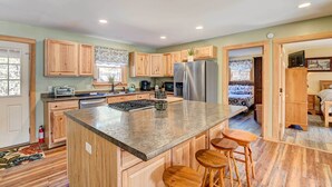 Une cuisine spacieuse avec des meubles en bois, un grand îlot central doté de tabourets, un réfrigérateur et un coin repas visible à travers une porte ouverte.